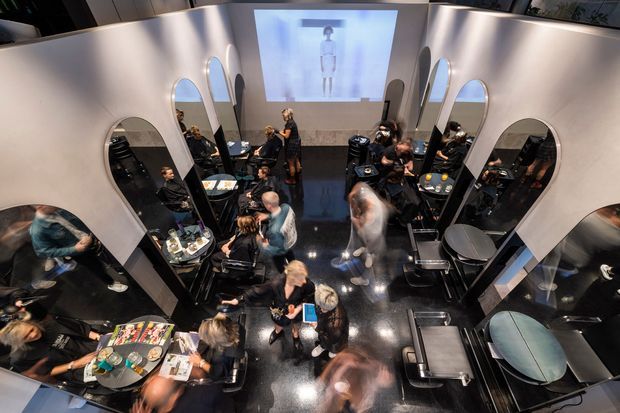 High-angle view of a dark, glossy commercial interior arranged like a hair salon or beauty studio, with multiple styling stations, black chairs, work tables, large oval mirrors, and several people gathered throughout the space. Curved pale walls and a large projected screen create a contemporary, theatrical atmosphere.