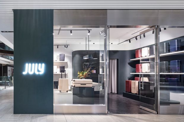 A contemporary retail showroom viewed from a mall or commercial corridor. The storefront has a dark teal sign panel with illuminated lettering, extensive glass doors and partitions, polished light flooring, black display shelving, track lighting, and a curated arrangement of boxed and appliance-like merchandise. The interior uses dark charcoal walls and shelving contrasted with white surfaces and metallic framing.