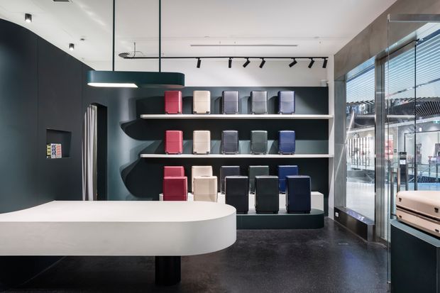 A contemporary retail showroom interior with dark charcoal walls, black flooring, and built-in display shelving presenting rows of compact colorful products. A large white rounded counter dominates the foreground, with pendant and track lighting overhead. Full-height glazed frontage with horizontal blinds opens views to the adjacent commercial environment.