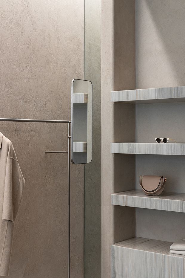 Close-up of a refined bathroom dressing and storage area, featuring textured taupe plaster walls, a slim dark-framed vertical mirror, dark metal towel rail with a hanging robe, and open built-in shelving in pale vertically grained timber or wood-look finish. Accessories include sunglasses, a handbag, and folded towels.
