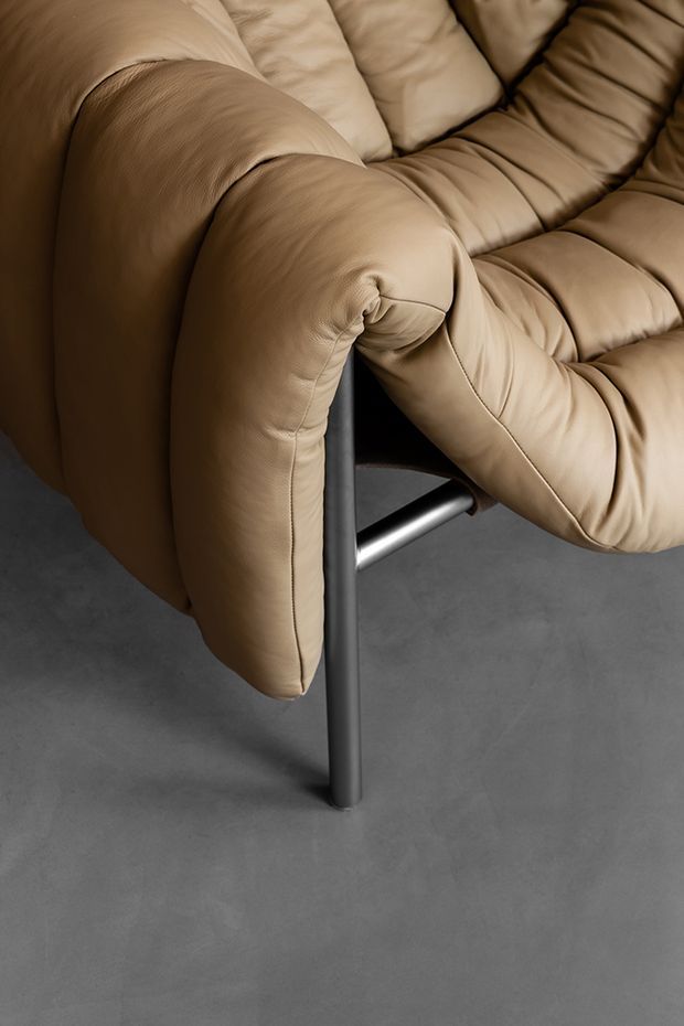 Close-up product-style image of a plush tan leather lounge chair with heavily padded, quilted upholstery and rounded armrests, supported by a dark metal tubular frame. The chair sits on a smooth grey concrete-like floor against a neutral background.