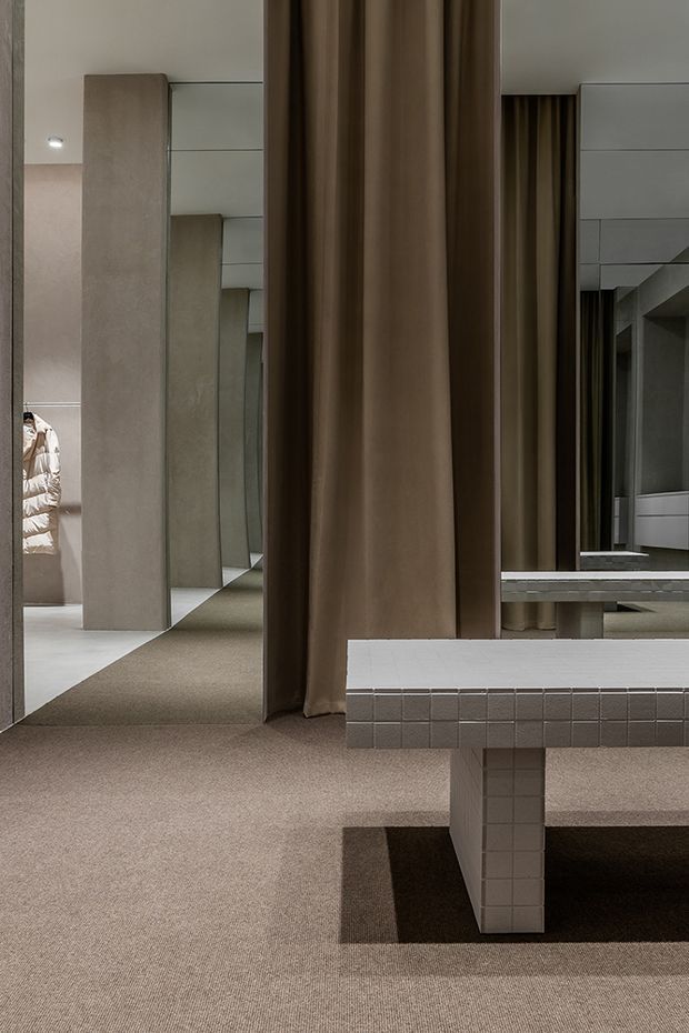A moody, minimalist interior featuring a tiled rectangular table or island in the foreground, set against warm brown floor-to-ceiling curtains, large mirrors, taupe plastered columns, and muted carpet flooring. The space has strong geometric lines, reflective surfaces, and a refined contemporary fit-out.