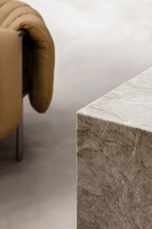 Close-up interior detail showing a rounded beige upholstered chair beside a substantial rectangular natural stone table or plinth with prominent veining and softly rounded edges. The setting has a warm, restrained, luxury aesthetic with a pale neutral wall and floor.