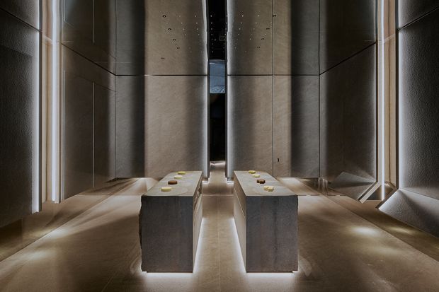A dark, highly refined contemporary interior corridor or display space with large grey stone or concrete wall panels, matching monolithic rectangular plinths, and integrated linear lighting at the floor and wall edges. The symmetrical composition creates a dramatic gallery-like atmosphere. Small objects are arranged on the central plinths, while a narrow opening and reflective surfaces extend the sense of depth.