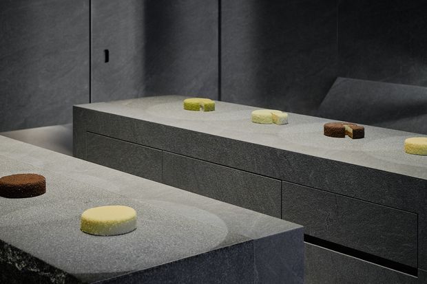 A moody, minimalist interior featuring large dark grey monolithic stone or concrete counters and integrated bench-like cabinetry. Several small round and rectangular food items are placed on the surfaces. The space has a restrained palette, sharp linear detailing, and a dark textured wall backdrop.