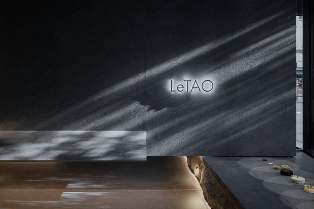 Dark, atmospheric contemporary interior featuring a large charcoal textured feature wall with the illuminated “LeTAO” sign. Strong diagonal sunlight creates dramatic shadow patterns across the wall and polished dark floor. A low stone-edged platform or counter occupies the foreground, with a glimpse of glazing and interior elements at the right edge.