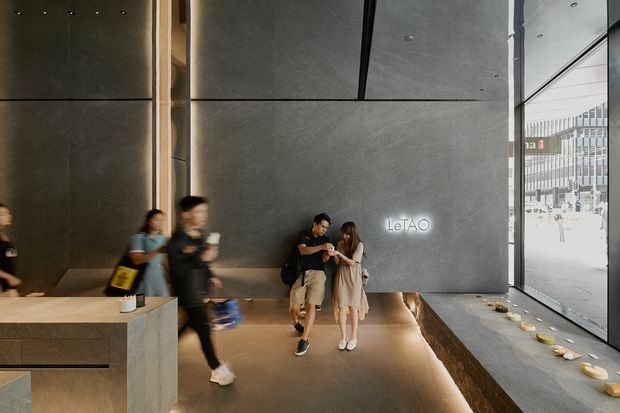 A contemporary commercial interior lobby or reception space with a dark, monolithic stone or concrete feature wall bearing illuminated “LeTAO” signage. A built-in stone reception counter occupies the foreground, while a long integrated bench runs along the wall and beside full-height glazing. Recessed ceiling lighting, warm under-seat illumination, and minimal detailing create a refined, atmospheric retail or hospitality setting. People are present, and an adjacent commercial building is visible through the glazing.