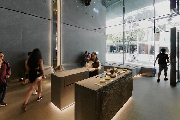 A contemporary commercial interior, likely a reception or hospitality space, with a large stone-clad central island and a long stone reception counter featuring integrated warm underlighting. Dark grey wall and ceiling panels create a monolithic architectural backdrop, while extensive floor-to-ceiling glazing opens the space visually to a bright urban plaza. Small plates and display items sit on the island, and several people move through the space.