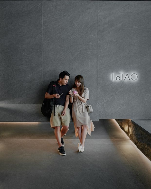 A modern commercial interior lobby or entrance with large dark grey textured wall panels, a polished grey floor, integrated linear lighting, and a glowing LeTAO sign. Two people are walking through the spacious, minimalist reception area. A recessed feature or planter-like opening is visible along the right side.
