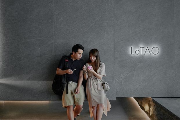 A dim, polished architectural interior featuring a large textured grey wall with illuminated “LeTAO” signage. Two visitors stand in front of the wall, beside a long recessed architectural feature with warm integrated lighting and a rough stone-like surface. The space has a refined, gallery or commercial hospitality feel with strong linear geometry and dramatic lighting.