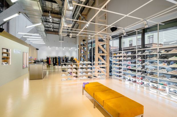Bright contemporary retail showroom with extensive wall-mounted and freestanding shoe displays, clothing racks, a central timber-framed display structure, exposed services, and long mustard-yellow upholstered benches. The space has polished pale flooring, large glazed frontage, and a high open ceiling with linear lighting and visible ductwork.