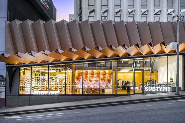 Street-level exterior view of a contemporary commercial retail storefront, with expansive glazed shopfronts, bright interior displays, and a dramatic sculptural folded-metal canopy projecting across the facade. The building is set along an urban street beside taller office or commercial buildings.