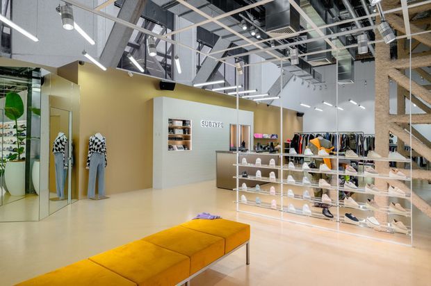 Bright contemporary fashion retail interior with clothing racks, glass display shelving for shoes and accessories, mannequins, a mustard upholstered bench, mirrored walls, and an exposed industrial-style ceiling with suspended linear lighting and visible services.