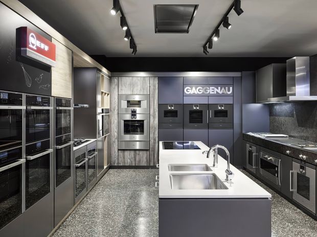 A sleek contemporary kitchen showroom displaying integrated ovens, induction cooking appliances, dark cabinetry, stainless-steel appliances, a large white island with double sinks and chrome fixtures, and stone-look benchtops. Track lighting and a dark ceiling create a polished commercial display environment.