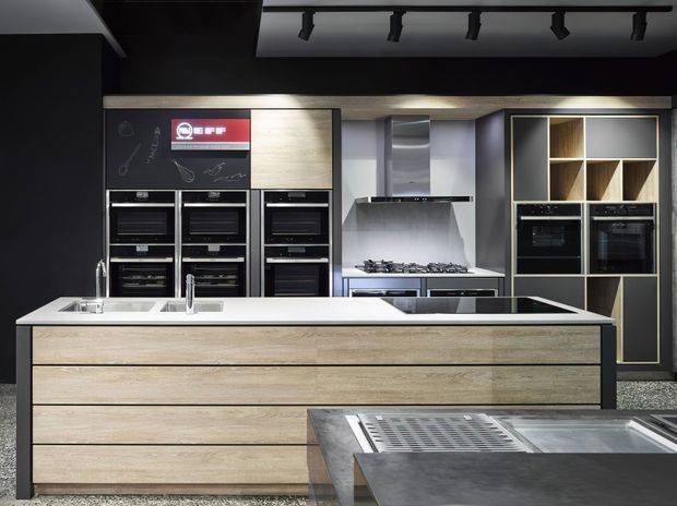 A high-end contemporary kitchen showroom featuring a large timber-fronted island with integrated double sinks and dark stone benchtops. The rear wall contains multiple built-in ovens, a gas cooktop with stainless-steel splashback and rangehood, and tall dark cabinetry with open timber display shelving. Black walls, ceiling track lighting, metallic appliances and layered work surfaces create a dramatic, sophisticated kitchen setting.