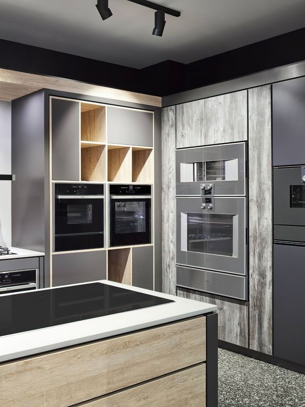 A contemporary kitchen featuring a large island with a glossy black cooktop and light timber-look drawer fronts. The rear wall combines integrated black and stainless-steel appliances, including multiple ovens and a refrigerator, with open shelving and cabinetry finished in weathered light wood and dark grey. The space has a dark painted upper wall, light ceiling, black track lighting, and a speckled hard-wearing floor.