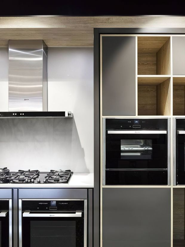 Close-up view of a contemporary kitchen featuring a gas cooktop, stainless-steel rangehood, white splashback and benchtop, and integrated black-glass ovens. Floor-to-ceiling cabinetry combines dark grey fronts with warm wood-grain open shelving and black-framed detailing.