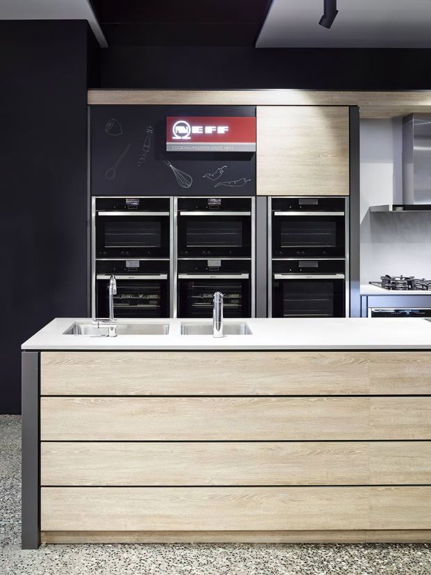 Contemporary showroom kitchen featuring a large central island with a white benchtop, integrated double sinks and chrome mixer taps. The island has light timber-look drawer cabinetry with dark side panels. Behind it is a bank of black built-in ovens, light timber cabinetry, dark wall panels, and a stainless-steel cooktop area. The space has a polished terrazzo floor and strong black, white, and pale timber contrast.