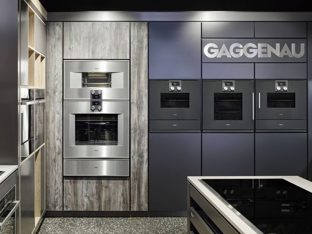 A high-end contemporary kitchen display featuring integrated Gaggenau ovens and appliances set within dark matte cabinetry. The room combines charcoal cabinetry, vertically grained timber-look wall panels, stainless-steel appliance fronts, a black reflective island benchtop with a light edge, and a terrazzo-style floor.