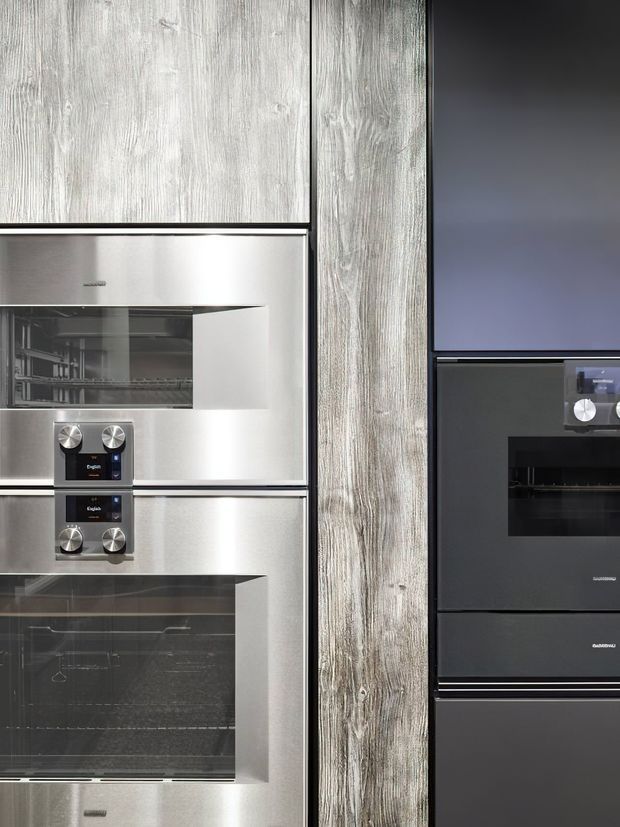 Close-up view of a contemporary built-in kitchen appliance wall featuring stacked stainless-steel ovens and a coffee machine on the left, contrasting with dark integrated ovens on the right. Vertical grey-toned wood-grain cabinetry or wall panelling separates the appliance banks, with sleek flush-fitting detailing throughout.