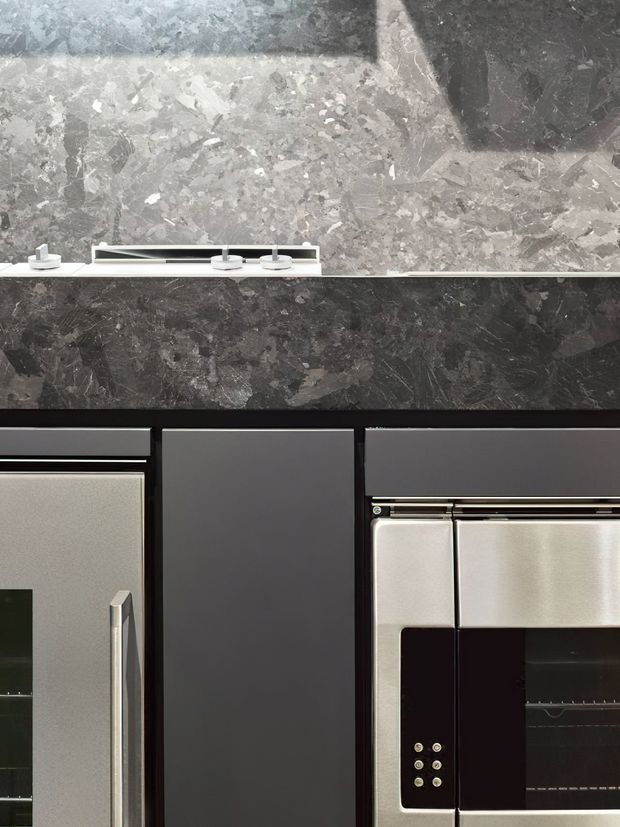 Close-up view of a sophisticated dark kitchen or bar-style joinery installation, featuring black and charcoal cabinetry, integrated stainless-steel appliances, and a dramatic grey veined stone backsplash and benchtop. A slim linear light or fixture sits along the stone surface, creating a refined, high-contrast contemporary look.