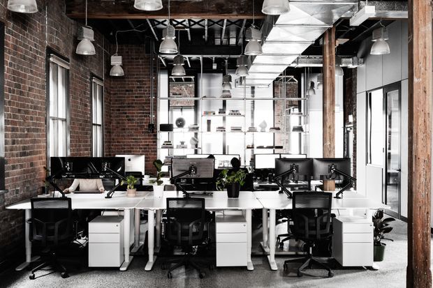 Open-plan commercial office with rows of white workstations, black ergonomic office chairs, computer monitors, shelving, and indoor plants. The space features exposed red brick walls, timber structural posts, visible black ceiling structure and services, extensive industrial-style pendant lighting, and large windows with abundant natural light.