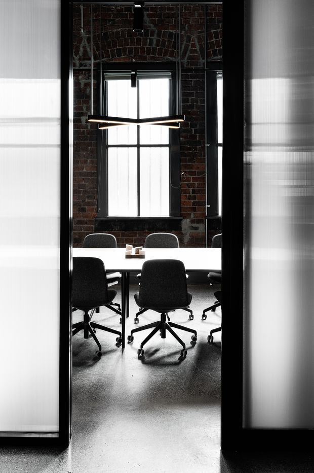 A narrow view through a dark-framed doorway into a compact industrial-style meeting room. A white rectangular conference table is surrounded by black and grey swivel office chairs, with a linear black pendant light suspended above. The room has exposed dark red brick walls, black-framed industrial windows with sheer blinds, polished grey concrete flooring, and reflective translucent panels framing the entrance. The image is high-contrast and monochromatic with warm brick accents.