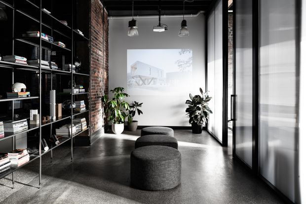 A dark, industrial-style interior lounge or reception space with exposed red brick walls, polished concrete flooring, black metal-framed glazing, and full-height open shelving filled with books and objects. Three rounded charcoal upholstered ottomans sit in the centre, with potted plants beneath hanging pendant lights. A projected architectural image appears on the white rear wall.