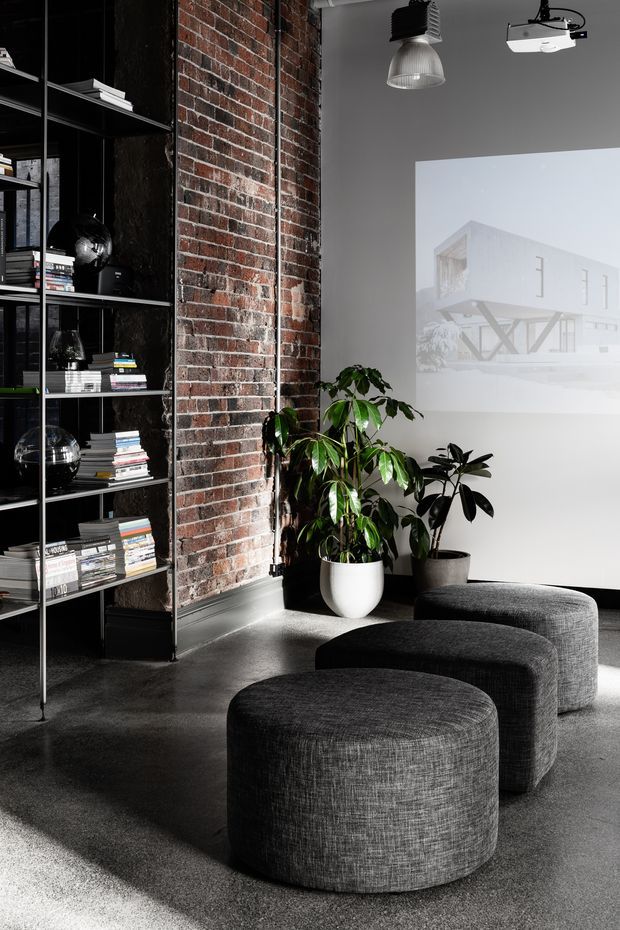 A moody, industrial-style interior featuring a dark built-in shelving unit filled with books and objects, an exposed red-brick wall, charcoal carpet, sculptural upholstered ottomans, indoor plants, and a projected architectural image on a pale wall. Exposed ceiling-mounted lighting and projector equipment contribute to the studio or presentation-space character.