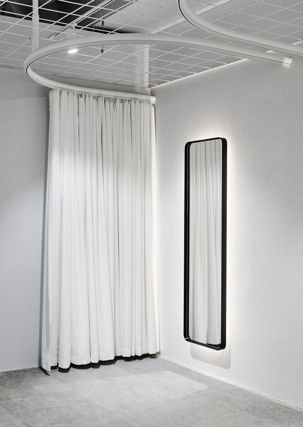 A minimalist monochrome interior corner with floor-to-ceiling white curtains, a tall rounded-rectangle black-framed mirror, pale textured walls, and grey carpet flooring. A large sculptural circular ceiling light is suspended beneath a visible white grid ceiling, creating a clean showroom or commercial display atmosphere.