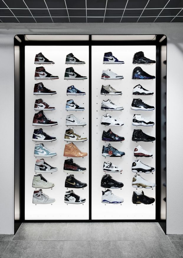 A retail display wall showcasing a large collection of sneakers arranged in four vertical columns on illuminated white panels within a black-framed recessed cabinet. The space has a dark grid ceiling, white walls, and large grey floor tiles, creating a clean, monochrome, high-contrast commercial setting.