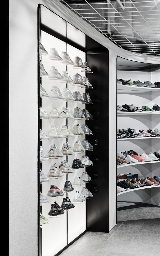 A monochrome, high-contrast retail shoe store interior with rows of sneakers displayed on illuminated wall shelving and curved white display units. The space features black-framed illuminated panels, dark accents, grey tiled flooring, and an exposed white grid ceiling with visible services.
