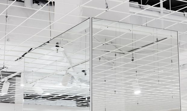 A close-up view of a commercial interior ceiling with a white exposed grid system, suspended lighting and services, and large clear glass partitions creating layered reflections. The composition is predominantly white and bright, with a minimal, open-plan architectural character.