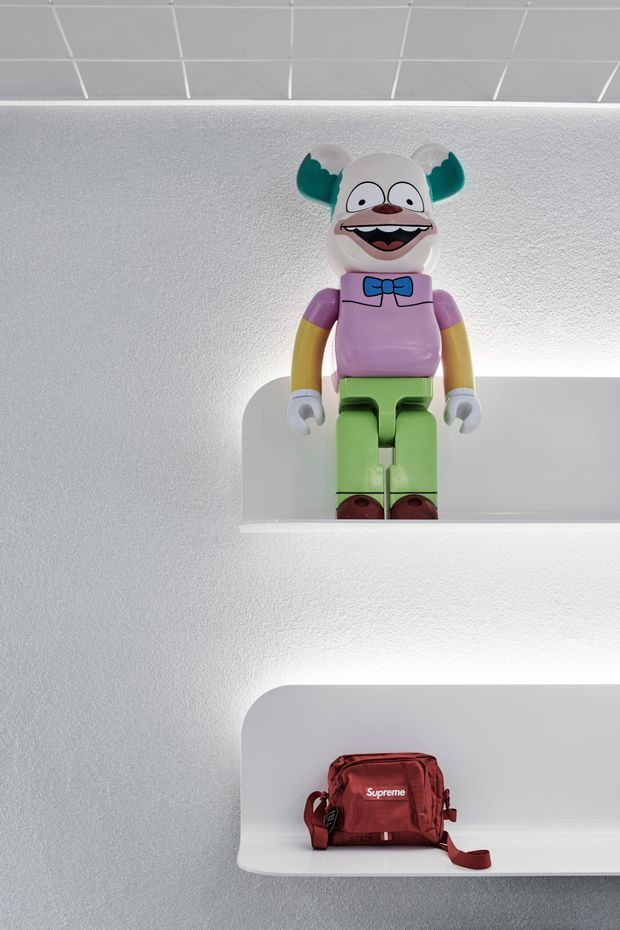 A minimalist display wall with two white, rounded-edge recessed shelves illuminated from above and below. The upper shelf holds a colourful cartoon-style toy figure, while the lower shelf displays a small red bag. The surrounding wall and shelves are predominantly white with a clean, contemporary retail or interior display aesthetic.