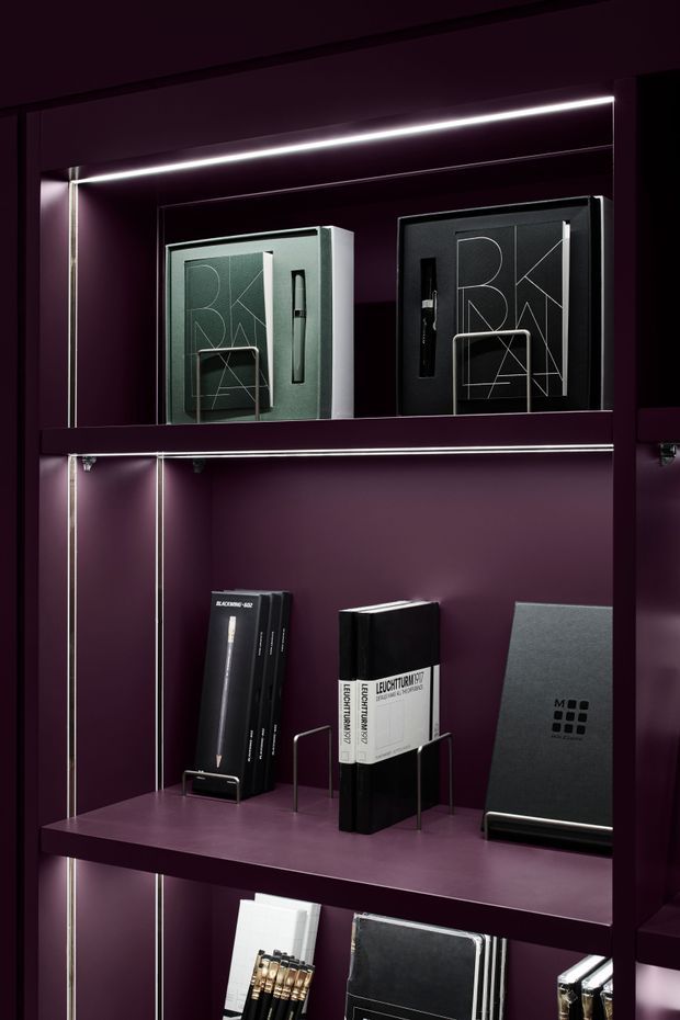 Close-up of a dark plum-purple built-in display shelving unit with integrated linear LED lighting and reflective backing. The shelves present monochrome design books, boxed materials, and graphic sample displays held in minimalist metal stands. The composition has a refined, dramatic showroom or retail-display character.