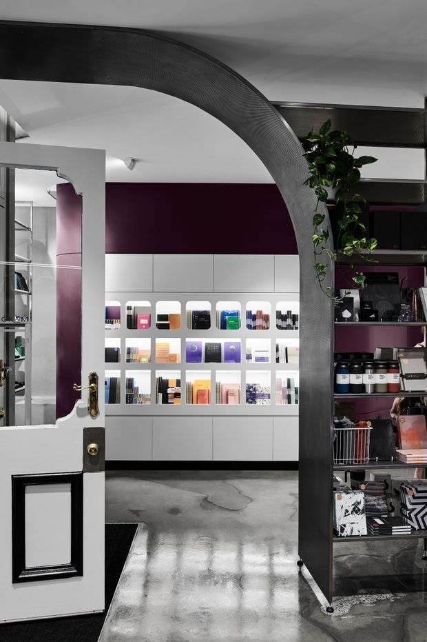 A dramatic retail or showroom interior viewed through an open, partially glazed white door. The space features a large curved dark ribbed architectural arch, polished concrete flooring, deep plum wall panels, white built-in display cabinetry, and illuminated shelves presenting books, samples, or merchandise. Dark open shelving on the right holds additional products and objects, while a trailing plant adds greenery. The overall composition is graphic, sculptural, and high-contrast.