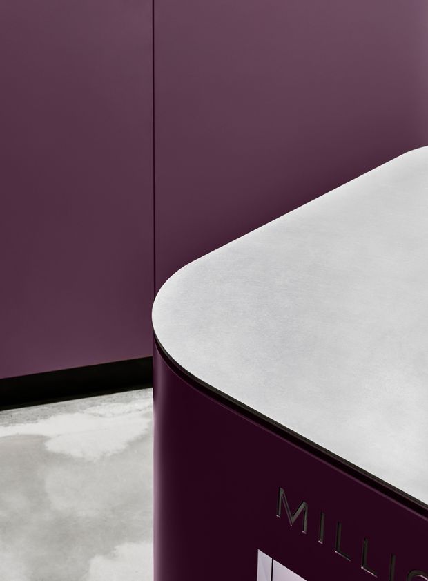 Close-up architectural detail of a deep burgundy curved built-in counter or reception unit with a pale grey rounded benchtop. The unit meets a matching burgundy wall with a dark skirting or base trim, while a mottled concrete floor is visible below. Partial metallic lettering appears on the front, suggesting a branded commercial interior or display counter. The image is tightly framed and focuses on the joinery, curved profile, and contrasting materials and colours.