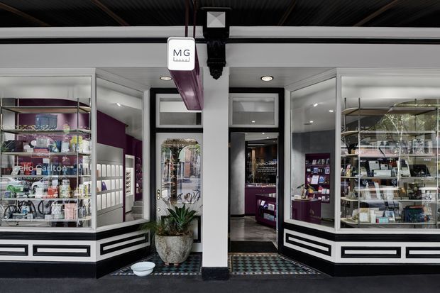 A contemporary retail storefront viewed from the exterior, with symmetrical glass display windows, white and black geometric trim, illuminated shelving, and purple-accented merchandise displays. The open central entrance leads into a compact boutique interior with patterned tile flooring and recessed ceiling lights.