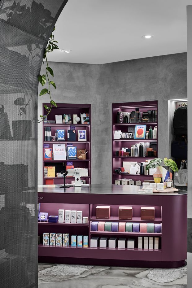 A contemporary retail shop interior with a deep burgundy-purple service and display counter, matching recessed wall shelving, and a grey mottled concrete-look wall finish. The counter displays small packaged goods and stationery-like products, while clothing racks are visible to the right. A curved perforated metal screen with trailing greenery partially separates the foreground from the retail area. The floor has an irregular polished concrete or stone-like finish, and recessed ceiling lights provide even illumination.