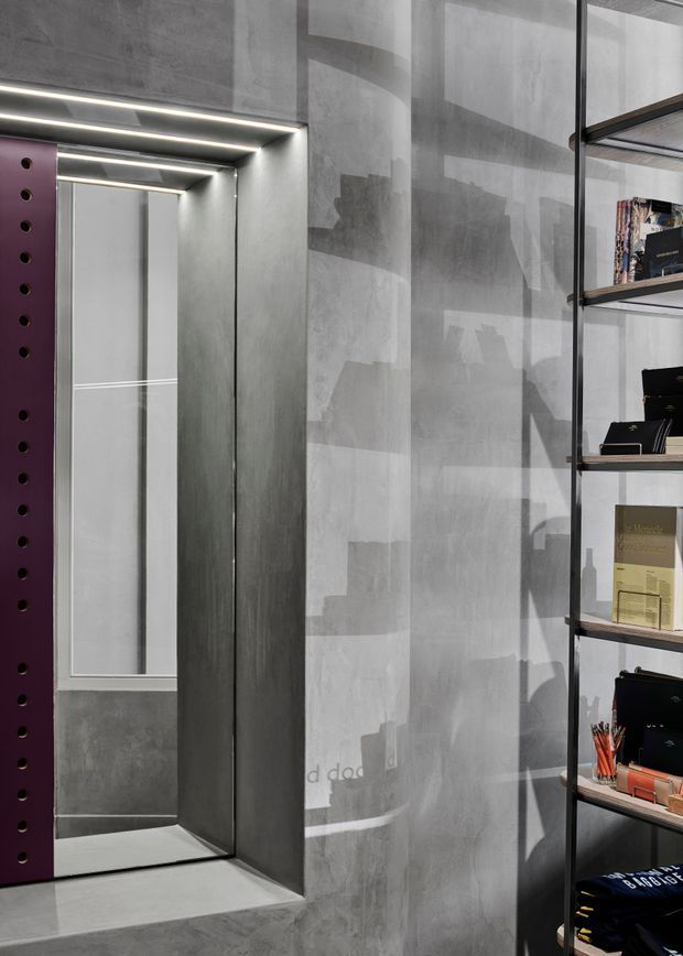 A narrow, modern retail or display interior with raw grey concrete-like wall finishes, a recessed illuminated doorway or fitting-room opening, and black metal-framed open shelving displaying books, bags, stationery, and clothing. Strong linear lighting and dark detailing create an industrial, monochromatic atmosphere.
