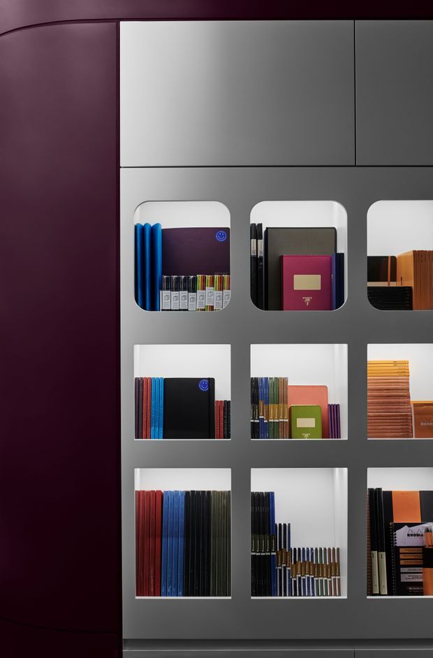 A close-up interior view of a contemporary built-in display shelving unit. The dark grey panelled joinery contains a grid of illuminated recessed compartments, some with rounded corners, neatly displaying colourful books, notebooks, folders, and sample materials. A deep burgundy wall or side panel frames the display, creating a refined, moody presentation.
