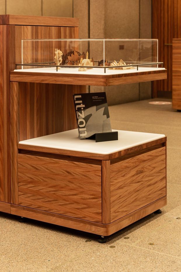 A warm, wood-paneled commercial exhibition or museum display area featuring a custom timber display cabinet on casters. The cabinet has a white solid-surface top, framed wood construction, a glass vitr ine case above containing a miniature architectural model, and an information booklet on the lower surface. The surrounding space includes matching timber cabinetry, dark vertical wall panels, and a neutral polished floor.