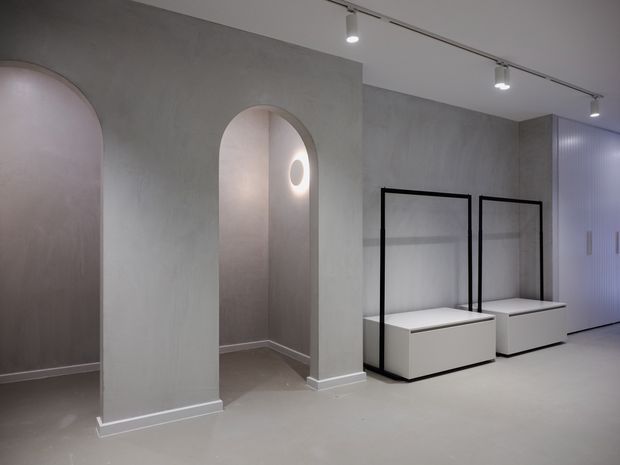 A minimalist, gallery-like interior with smooth light-grey plaster walls, rounded arched openings, pale flooring, and streamlined white display benches framed in black metal. Track lighting and a softly glowing circular wall light create a calm, refined presentation space, with tall ribbed cabinetry along the right wall.