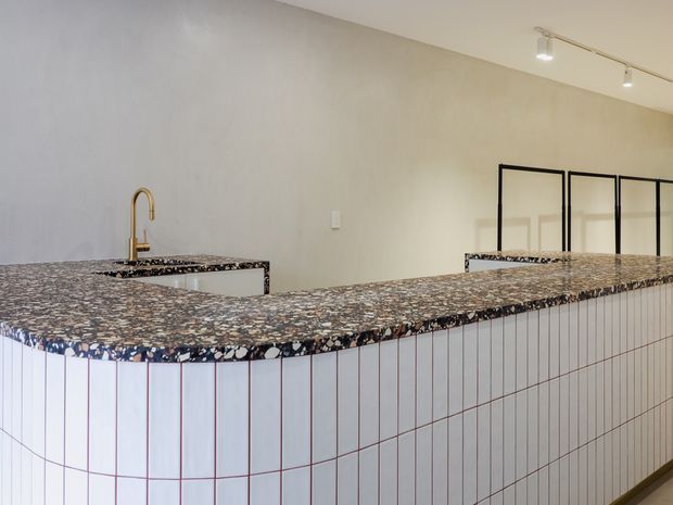 A contemporary kitchen or hospitality-style counter with a large L-shaped tiled island or bar. The counter has a dark, heavily patterned stone or terrazzo-like benchtop, while the curved-front base is clad in vertically oriented pale tiles with dark grout and subtle pinkish lines. A brass gooseneck tap, inset sink, black-framed internal glazing or screens, and ceiling-mounted track lighting are visible. The space is clean and minimally furnished.