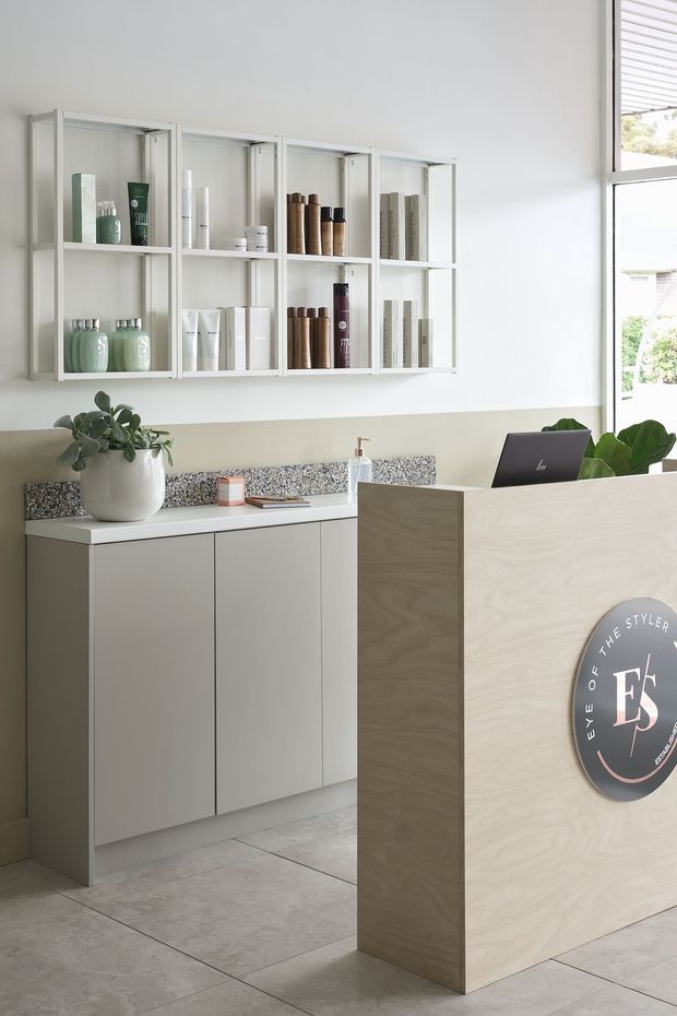 A bright, minimalist reception area, likely in a beauty salon or styling studio. The space features a pale timber reception desk with a circular logo, white built-in cabinetry, open wall shelving displaying beauty products, a speckled stone-look benchtop, large light-grey floor tiles, indoor plants, and a laptop. Natural light enters through a tall window with horizontal blinds.
