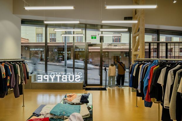 A contemporary clothing retail shop viewed from inside, with garment racks lining both sides, folded clothes on a central display table, polished light flooring, suspended linear fluorescent lighting, and full-height glazed storefront doors and windows opening onto a commercial street. A prominent sculptural timber-like column rises near the entrance.