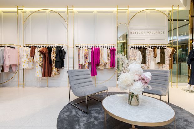 A refined high-end fashion boutique interior with white decorative wall panelling, illuminated display niches, and elegant gold-toned clothing racks. Garments are arranged along the walls, while two grey upholstered lounge chairs and a round marble-topped coffee table sit on a dark circular rug in the foreground. A floral arrangement provides a soft decorative focal point, and mirrored glazing reflects the retail space.