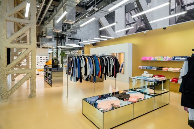 An expansive contemporary retail clothing store with exposed industrial services and structure, suspended linear lighting, polished concrete-style columns, and a mustard-yellow feature wall. Clothing racks, folded garments, and glass-topped display tables occupy the open showroom floor.
