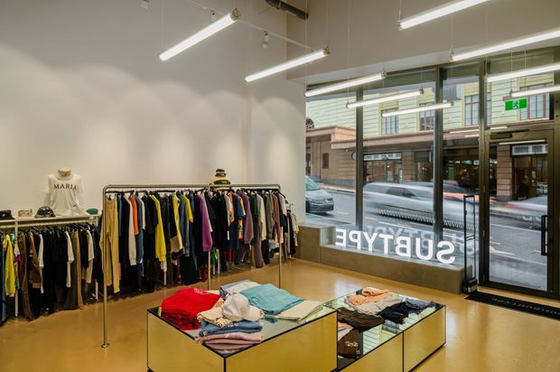 A bright contemporary clothing retail store interior with polished timber flooring, white walls, exposed ceiling services, linear pendant lighting, freestanding garment racks, folded clothing displays on mirrored tables, and full-height glazed frontage opening onto a city street.