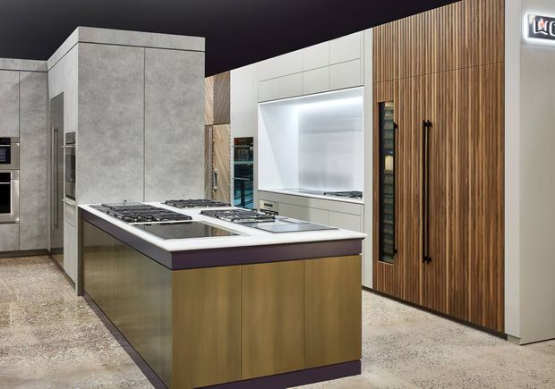 A contemporary kitchen showroom featuring a large central island with multiple gas cooktops, integrated appliances, extensive built-in cabinetry, a white tiled or panelled backsplash, and a tall timber-look storage wall. The space has polished concrete or terrazzo-style flooring, dark ceilings, and concealed LED lighting.